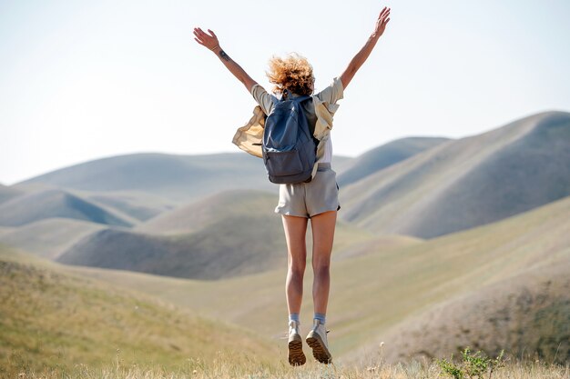 Photo euphoric woman jumping on the spot amidst a beautiful hills scenery. throwing hands in the air. from behind.