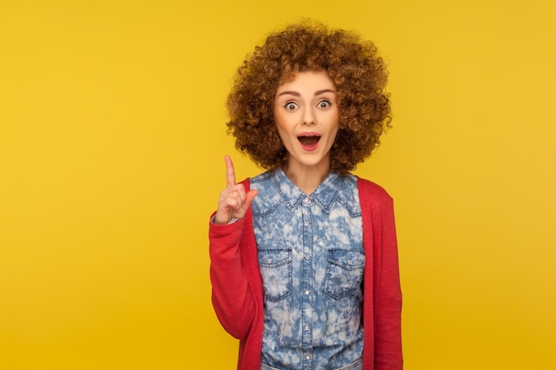 Eureka, I know answer. Portrait of amazed inspired woman with curly hair pointing finger up and having genius idea, surprised by sudden clever solution. studio shot isolated on yellow background