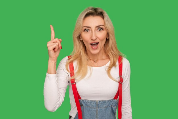 Eureka portret van geïnspireerde verbaasd volwassen vrouw in stijlvolle denim overalls wijzende vinger omhoog verrast door creatief idee geniale slimme oplossing indoor studio shot geïsoleerd op groene achtergrond