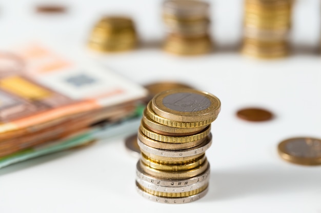 Euro coins standing in column on white table with heap of banknotes in background.