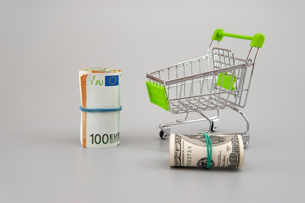 euro and dolar banknotes in rolls with supermarket trolley on gray background