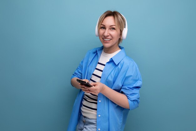 European adorable blond woman in shirt listening to music in wireless headphones on blue background