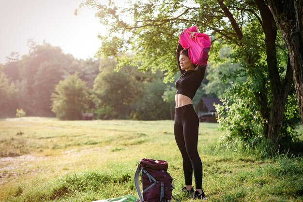 European athlete girl take off pink jacket on green meadow near\
forest. young beautiful smiling woman with closed eyes wear\
sportswear. concept of resting and tourism on nature. sunny\
daytime