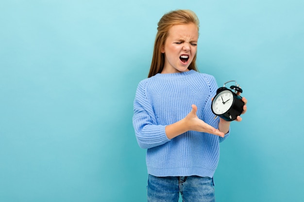 European attractive girl screams and holds an alarm clock on a light blue wall