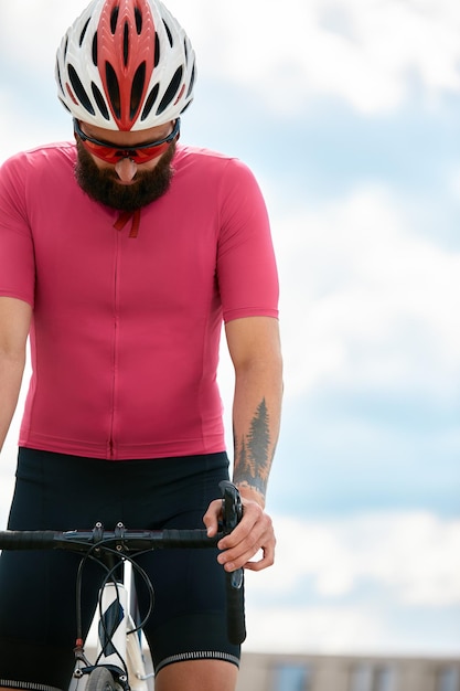 Foto ciclista europeo con la barba che indossa un casco con maglietta rosa consigliare alle persone di proteggere il cielo blu sullo sfondo