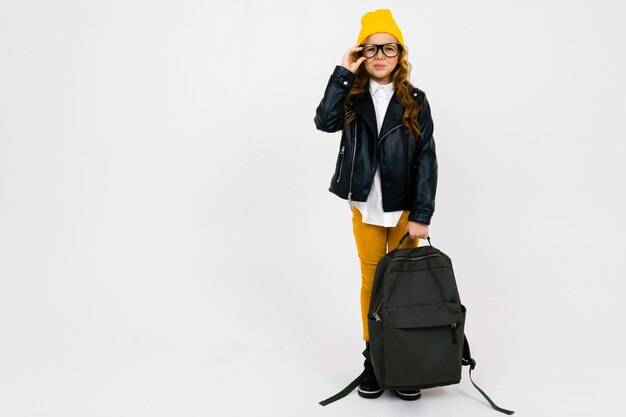 European beautiful girl dressed in a yellow hat, glasses and a leather jacket with a backpack in his hand in full growth on white