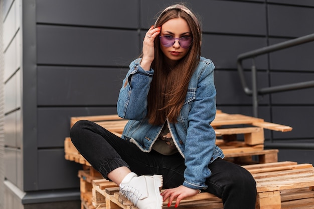 European beautiful young hipster woman in fashionable violet\
glasses in stylish denim jacket in black jeans posing sitting on\
vintage wooden pallets in the city. attractive girl n trendy wear\
outdoors