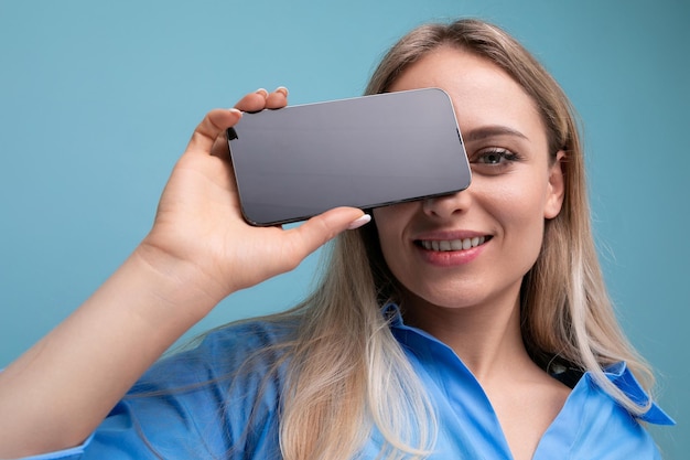 European blond girl holding smartphone screen with mockup for web page near her face on blue