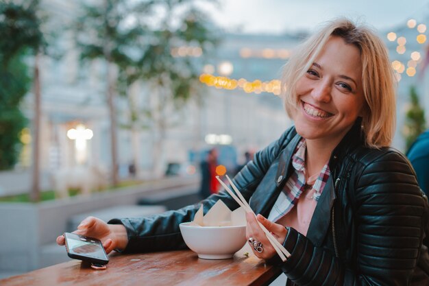 European blonde woman eating rice Pho Bo with wooden sticks table street Vietnamese cafe in Moscow.