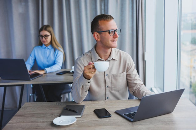 European businessman looking at something on laptop while working and drinking coffee The concept of a modern successful person Young guy in glasses sitting at the desk in the office with a laptop