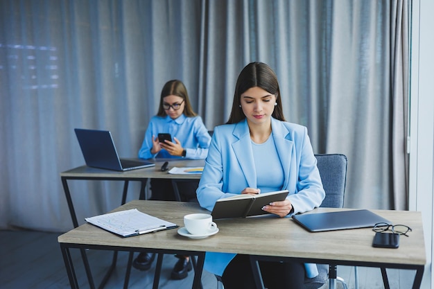 유럽 여성 사업가가 유럽 동료가 백그라운드에서 일하는 동안 휴대전화로 통화하고 있는 현대적인 성공적인 여성의 개념 햇살 가득한 사무실의 책상에 앉아 있는 어린 소녀들