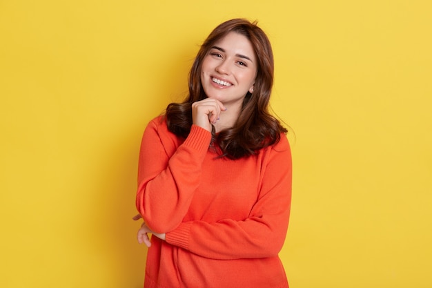 European charming girl with dark hair, holding fist under chin and smiling broadly, looking directly at camera, posing over yellow, expressing happiness.