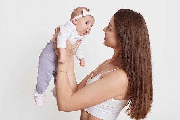 彼女の手で女の赤ちゃんを保持し、白い背景で隔離の愛を込めて彼女を見ているヨーロッパの陽気な美しい若い女性、弓でヘアバンドを身に着けているママと少女。