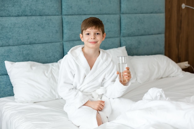 A European child holds a glass of clean water A boy in a white coat is sitting on the bed with a glass of water