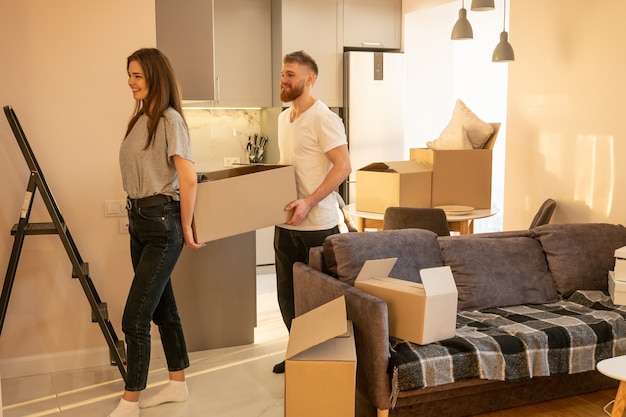 European couple carrying cardboard box with things at home. Concept of moving in new flat. Idea of young family. Beautiful girl and bearded man. Interior of studio apartment. Sunny daytime