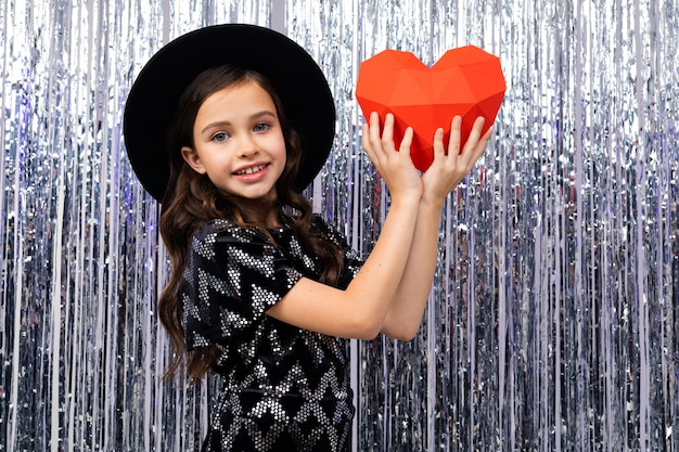 European cute girl in holiday dress and hat holds a red paper heart on a shiny