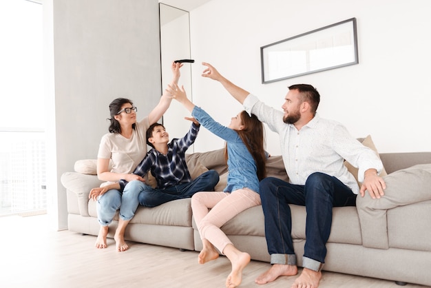 european family with son and daughter sitting on sofa together in living room at home, and trying to grab smartphone from woman