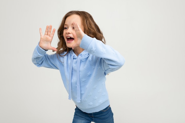 European girl in a blue hoodie shouts while holding hands at the mouth on a light wall