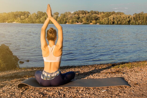 A European girl in a light tracksuit practices yoga on the river bank on a rubber yoga mat