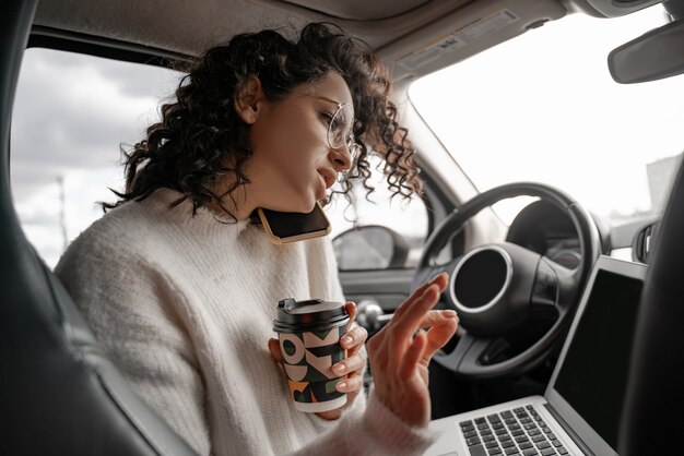 European girl talking on mobile phone on driver's seat in personal automobile. Focused curly businesswoman wearing glasses. Person holding laptop computer and coffee paper cup. Concept of multitasking