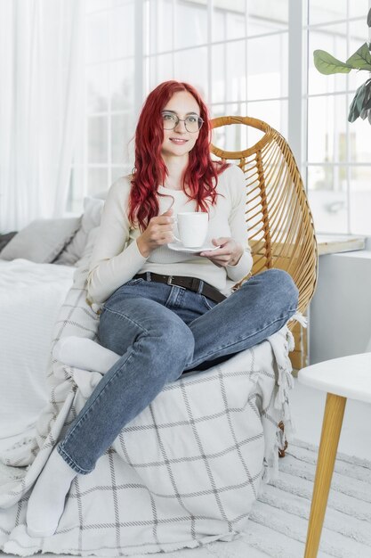 Foto una ragazza europea con i capelli rossi si siede in una giacca leggera su una sedia di vimini e tiene una tazza di caffè