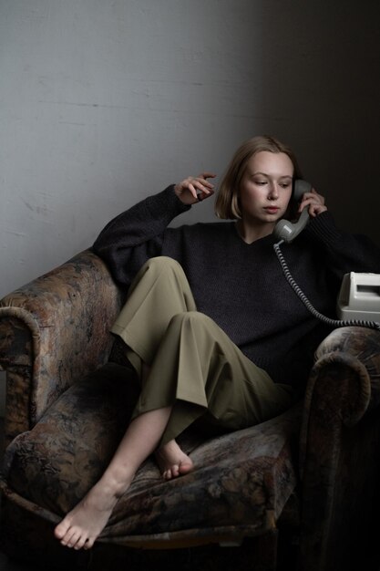 A European girl with short blonde hair in a knitted sweater poses with old phone for a catalog