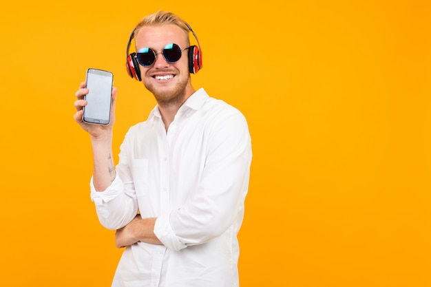 European guy in a white shirt and sunglasses listens to music in large headphones and shows the phone screen on yellow.