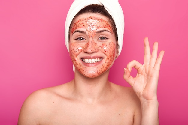 European lady with cleansing mask on face, shows okay sign, has toothy smile, wears white towel, has naked body