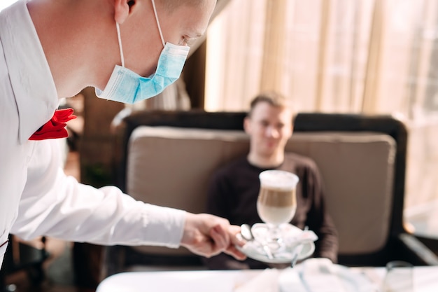 A European-looking waiter in a medical mask serves Latte coffee.