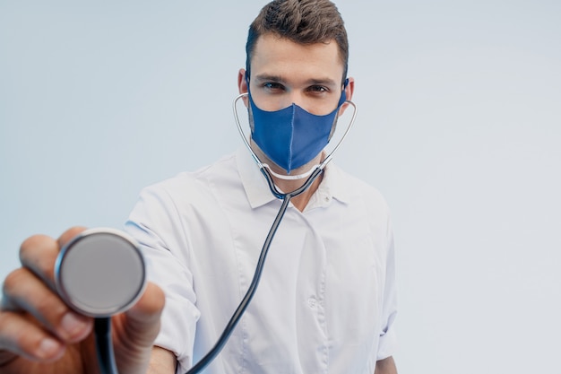 European male doctor in protective mask using stethoscope. Young man wearing white coat. Isolated on gray background with turquoise light. Studio shoot. Copy space.