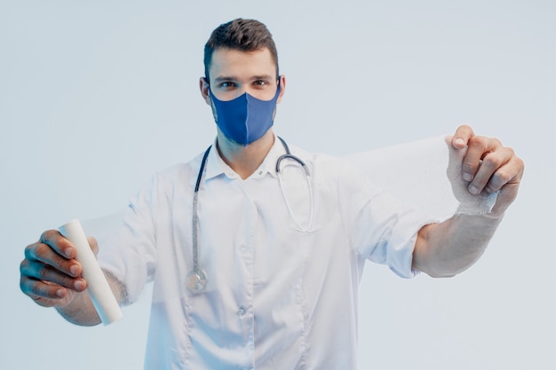 European male doctor showing medical bandage. Young man with stethoscope wearing white coat and protection mask. Isolated on gray background with turquoise light. Studio shoot. Copy space