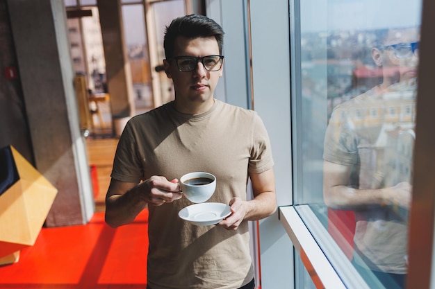 European man drinks coffee and looks out the window in a cafe A young smiling student in glasses stands near the window Recreation leisure and free time Modern male lifestyle