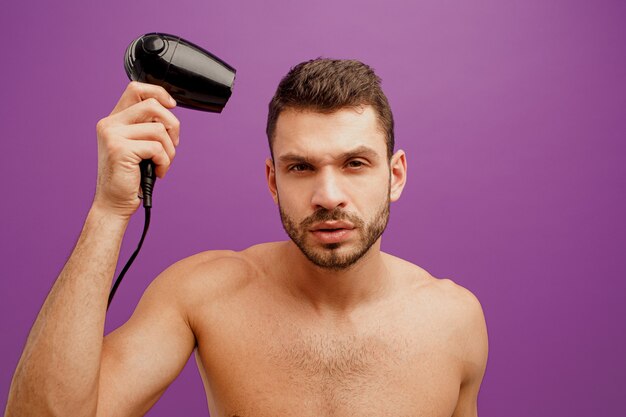 European man dry his hair with hairdryer. Focused handsome young bearded guy looking at camera. Isolated on purple background. Studio shoot. Copy space