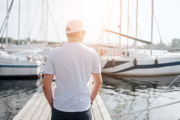 European man stand on pier and look at yacht. He keeps hands in pockets. Young man look to left. He is serious and calm