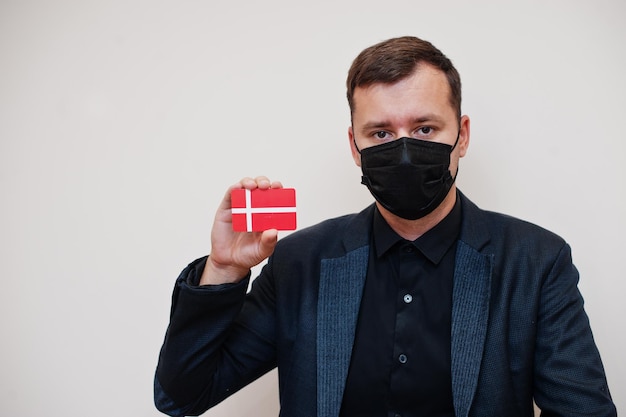 ヨーロッパ人男性は黒のフォーマルを着用し、フェイスマスクを保護します白い背景に分離されたデンマークの旗カードを保持しますヨーロッパコロナウイルスCovid国の概念