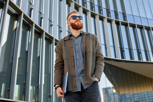 Европейский мужчина с красной бородой, одетый в рубашку и куртку, стоит у входа в бизнес-центр с ноутбуком