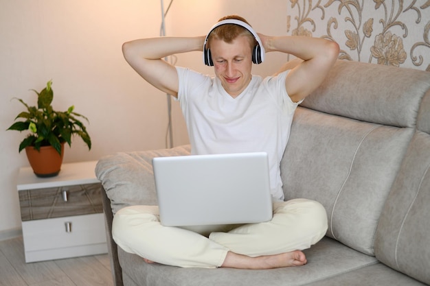 European man with throws his hands behind his head sits on the couch in headphones and looks at the laptop