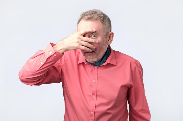 Uomo maturo europeo che si copre il viso a mano e fa capolino alla telecamera