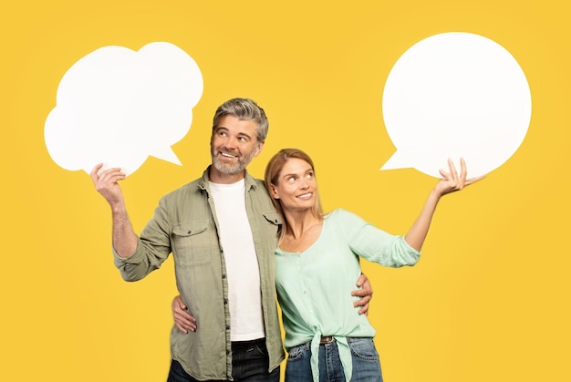 European middle aged couple holding empty speech bubbles and embracing posing on yellow background