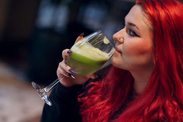 European plus size woman drink green smoothie in cafe young red pink haired body positive girl sitti...