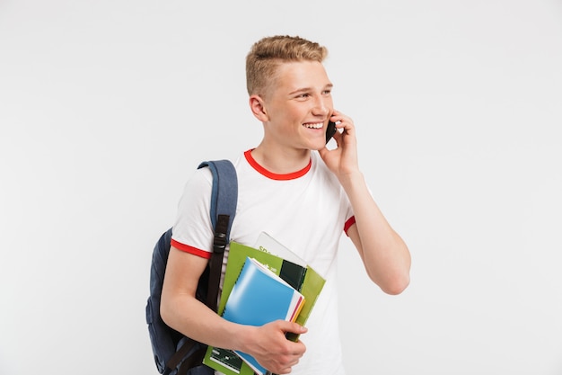 european positive student guy wearing backpack talking on smartphone with smile while holding colorful exercise books isolated on white