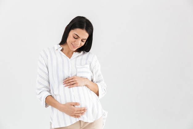 европейская беременная женщина улыбается и трогает свой большой живот, стоя изолированно над белой стеной