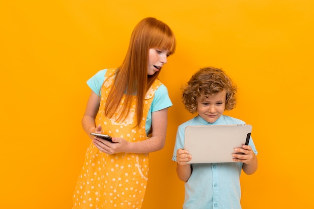European red-haired surprised boy and girl with phone and tablet isolated on yellow bright background.