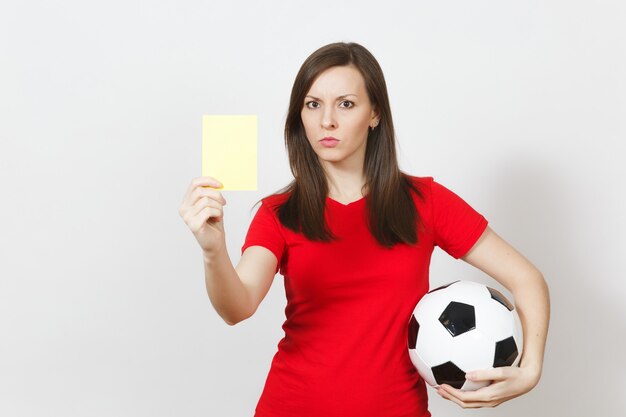 European serious severe young woman, football referee or player in red uniform showing yellow card, holding soccer ball isolated on white background. Sport play football, healthy lifestyle concept.