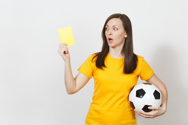 European serious severe young woman, football referee or player\
in yellow uniform showing yellow card, holding soccer ball isolated\
on white background. sport play football, healthy lifestyle\
concept.