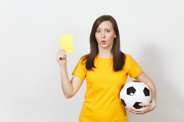 European serious severe young woman, football referee or player\
in yellow uniform showing yellow card, holding soccer ball isolated\
on white background. sport play football, healthy lifestyle\
concept.