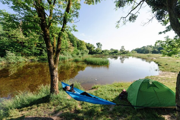 European sport girl lying on hammock near tent on green meadow\
near lake. young beautiful woman wear sportswear. concept of\
resting and tourism on nature. camping vacation. sunny summer\
day