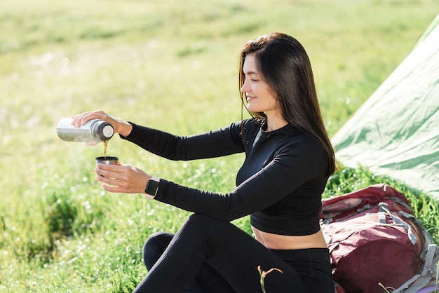 European sport girl sit on fitness mat near tent and drinking\
tea. green meadow. young pretty woman wear sportswear and enjoying\
sunlight. concept of resting and tourism on nature. camping\
vacation