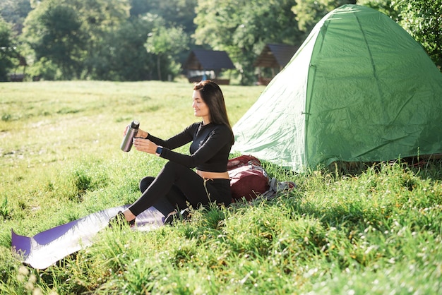 European sport girl sit on fitness mat near tent and drinking\
tea. green meadow. young pretty woman wear sportswear and enjoying\
sunlight. concept of resting and tourism on nature. camping\
vacation