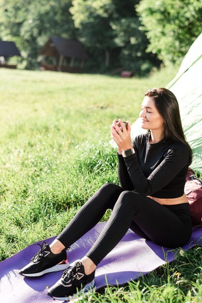 European sport girl sit on fitness mat near tent and drinking\
tea. green meadow. young pretty woman wear sportswear and enjoying\
sunlight. concept of resting and tourism on nature. camping\
vacation